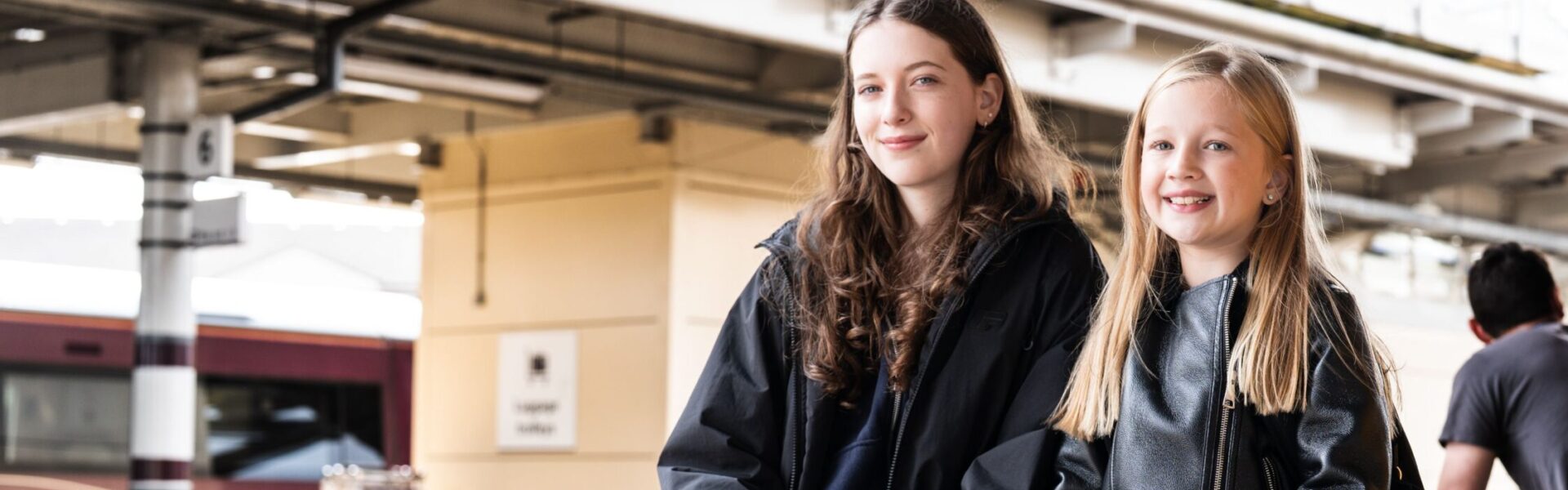Railway station platform with two young people seated side by side, with trains and station structures around them.