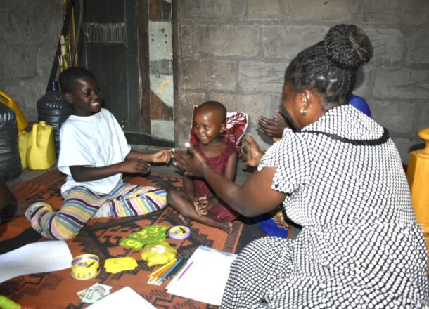 An adult support worker sits on a patterned mat with two children in a modest indoor setting, engaging in an interactive activity with educational toys, paper, and crayons. The group is gathered on the floor near a stone wall and wooden door, with everyday household items in the background. The scene reflects Railway Children’s community outreach work providing safe spaces and support for vulnerable children.