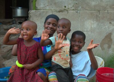 A mother sits with her three smiling children outside their home, all waving happily, reflecting family strength and the supportive work Railway Children does with vulnerable families in East Africa.