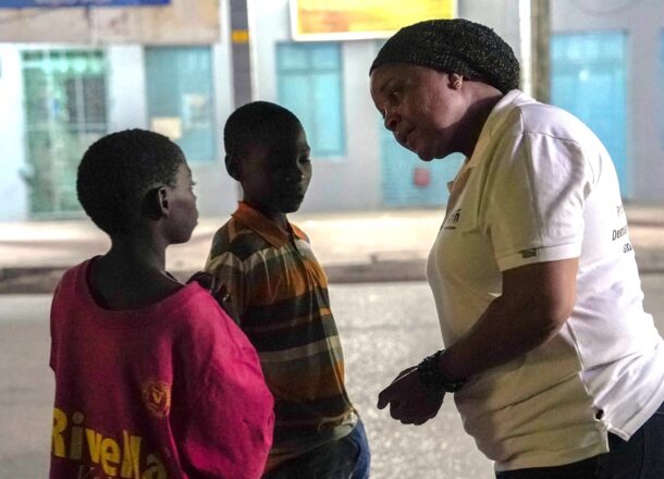 Adult wearing a white shirt speaking with two children standing on a street at night, with buildings and shopfronts visible in the background.