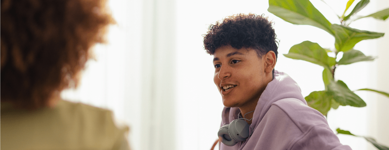 A young person wearing a lavender hoodie and headphones sits in a bright, modern room, smiling while engaged in a supportive conversation with another individual. A large green plant adds a calm, welcoming atmosphere in the background.