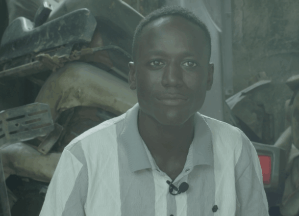 A young person wearing a collared shirt and a small clip‑on microphone is seated in a workshop‑style environment with stacked vehicle parts and equipment behind him. The setting reflects Railway Children’s support programmes that help young people access skills training and safer opportunities.
