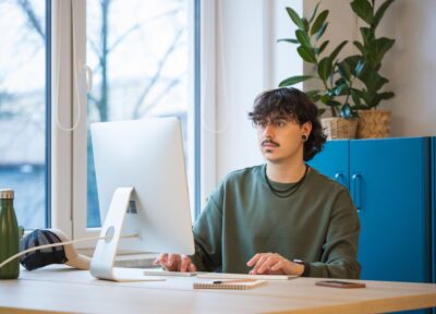 A young person sitting at a desk working on a computer in a bright, modern office with plants and natural light from a large window.