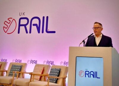 A man standing behind a podium giving a speech in front of a wall which reads UK RAIL.