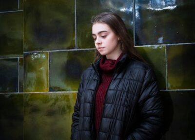 A young person with dark hair and wearing dark clothing stood in front of a green tiled wall looking down.