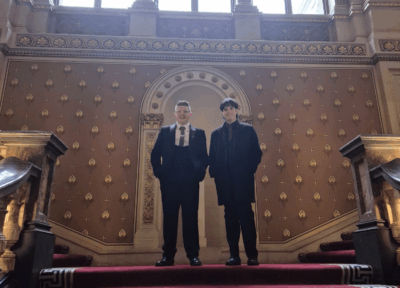 Two teenage boys wearing smart suits stand half way up a grand staircase, facing the camera.