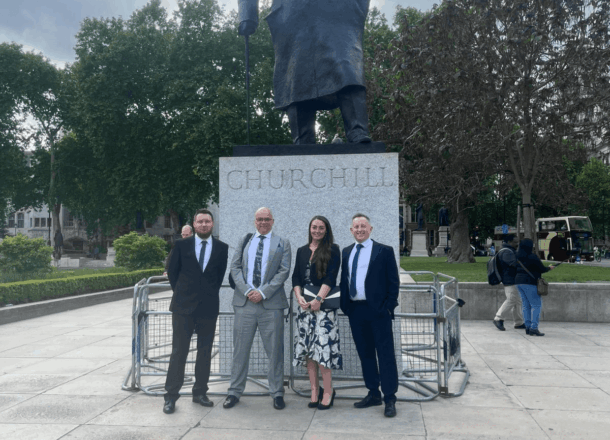 Four adults dressed smartly stand below a large statue, facing the camera.
