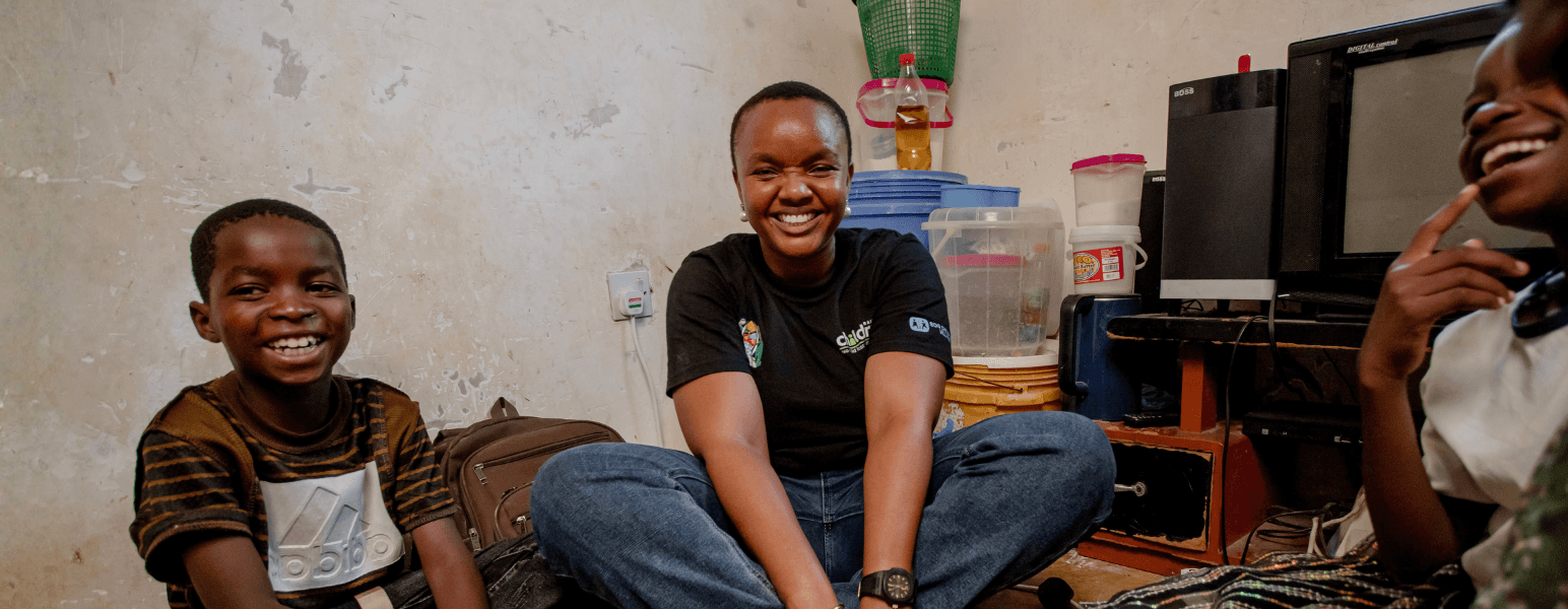 A Railway Children Africa project worker sitting on the floor of a family home with some children laughing and smiling whilst playing a game.