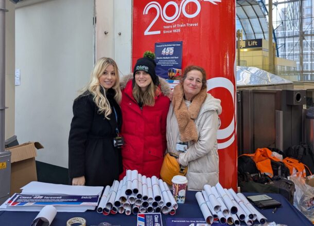 Three people at a railway station information stand with leaflets and merchandise beside a “200 Years of Train Travel” display.