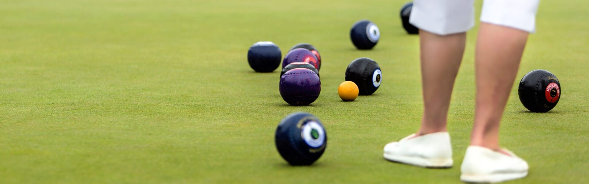 Image of a bowls pitch with various coloured balls and the bottom half of a bowls player wearing white cropped trousers and white shoes.