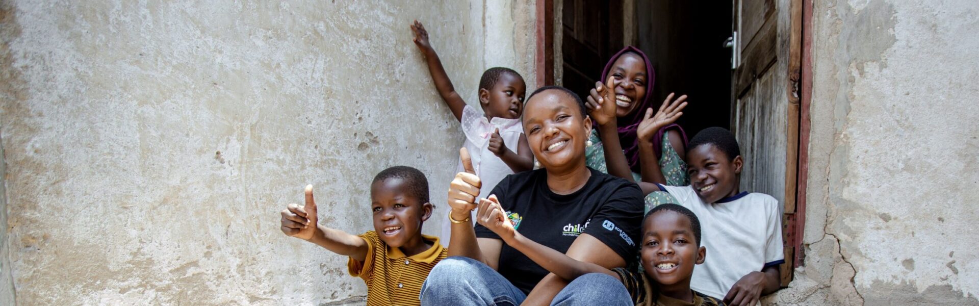 A Railway Children Africa project worker sat on a step outside a family home with a happy family raising their arms in the air.
