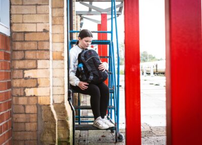 A young teenage girl sat on the steps next to a train station platform holding a large rucksack.