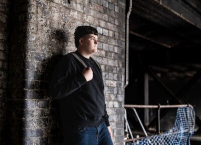 A teenager stood against a brick wall holding a backpack.