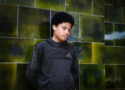 A teenager in the UK against a dark green tiled wall wearing a dark coloured sports jacket and looking down at the floor.