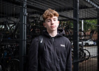 A young person in the UK wearing dark coloured clothing in front of a bike shed.