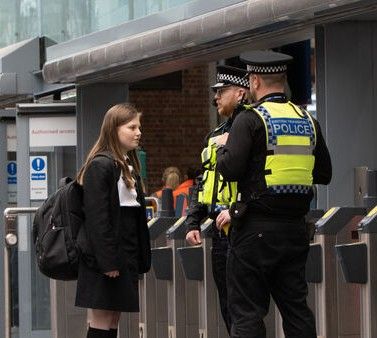 British Transport Police officers talking to a teenage girl in school uniform at a train station in the UK.