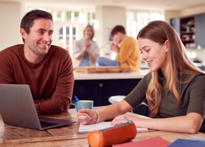 A father in front of a laptop sat at a kitchen table with his teenage daughter and talking, in the background there are other members of the family.
