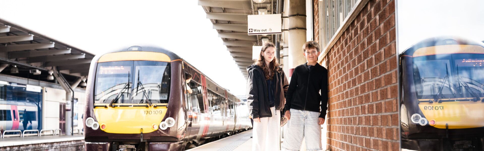 Two young people stood on a railway station platform in the UK smiling together with a train next to them.