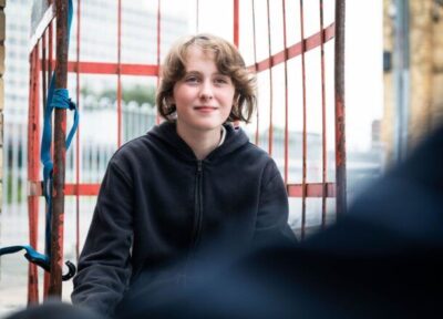 A young person sat in front of a fence on a train station platform.