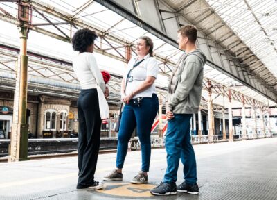 A rail industry worker providing support to two young people at a train station in the UK on a platform.