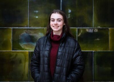 A young person wearing dark clothing stood against a green tiled wall and smiling at the camera with their hands in their pockets.