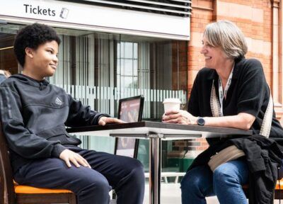 A teenager sat at a table in a cafe at a train station talking to a Railway Children youth practitioner.