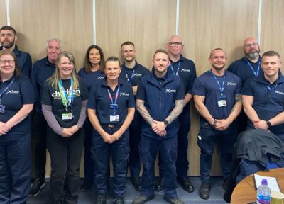 A group of adults wearing navy ScotRail uniforms stand together in an office meeting room with glass walls with a Railway Children staff member. The group is posing for a photograph, with desks, chairs and refreshments visible in the foreground.