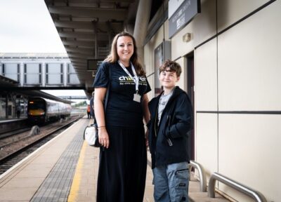Railway Children youth practitioner with a young teenager on a railway station platform beside an incoming train, with station infrastructure visible in the background.