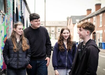 Five young people talking together on a residential street beside a colourful mural.