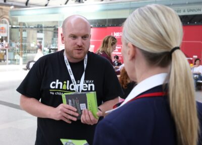 A Railway Children member of staff wearing a black branded T-shirt and lanyard holding awareness materials and speaking to a rail industry worker at a station in the UK with LNER boards behind them and passengers.