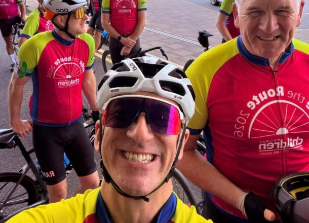 A group of cyclists wearing matching pink and green Ride the Route 2026 charity cycling jerseys gather with their bikes outside a building during a rest stop. One cyclist wearing a white helmet takes a selfie in the foreground, while other riders stand nearby with bicycles. Several vans and parked vehicles are visible in the background beneath an overcast sky.