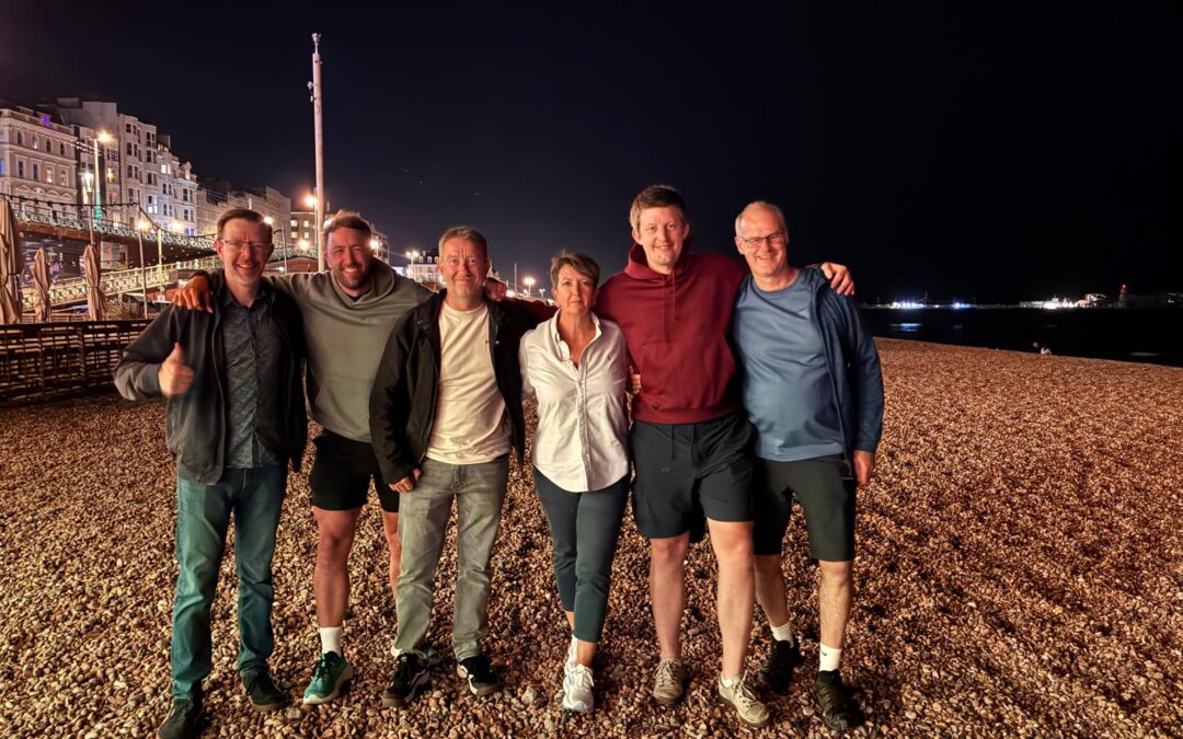 Supporters pose on Brighton beach at night following a Ride the Route charity cycling challenge, with the illuminated seafront and coastline in the background.