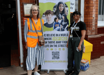 Two people stand beside a Railway Children banner outside Lichfield City railway station during a community engagement event. One person wears a high-visibility rail vest and the other wears a Railway Children branded t-shirt.