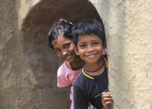 Two children standing in a doorway, looking towards the camera, with a textured wall and archway in the background.
