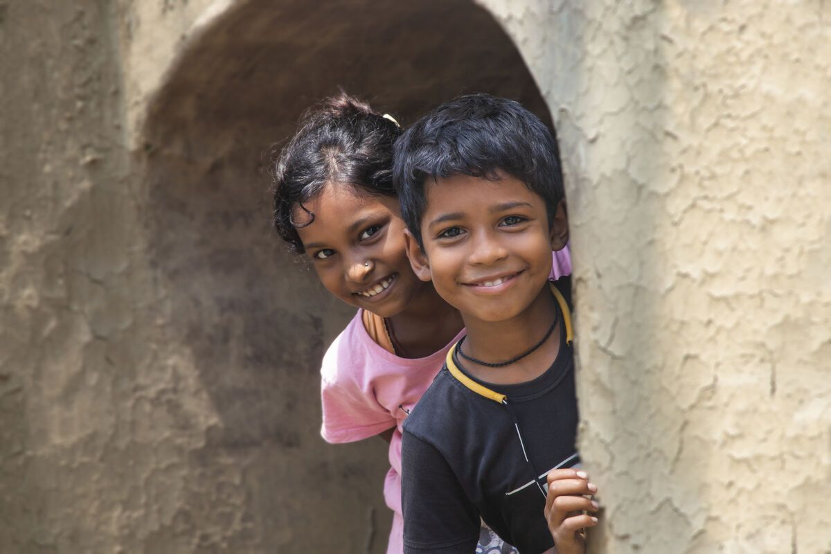 Two children standing in a doorway, looking towards the camera, with a textured wall and archway in the background.