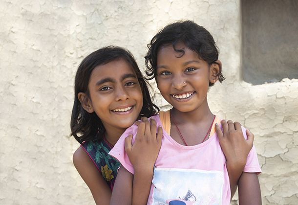 Two Indian children standing together and smiling outdoors in front of a textured wall, representing community, childhood wellbeing and education support.