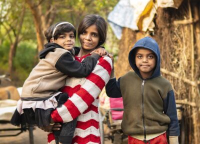 Indian adult holding their child with another child standing nearby in an outdoor community setting, highlighting family support, child wellbeing and community development.