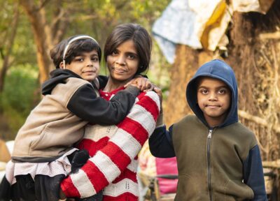 Indian adult holding their child with another child standing nearby in an outdoor community setting, highlighting family support, child wellbeing and community development.