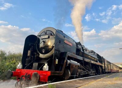 Steam locomotive 70000 Britannia stands at a railway station platform under a blue sky with scattered clouds. White steam rises from the locomotive, which is coupled to a set of passenger carriages.