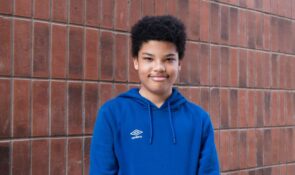 A young person in a blue hoodie standing in front of a red brick wall and looking towards the camera.