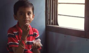 A child in India wearing a red striped top sitting beside the window of a train carriage, holding a small bag of food.