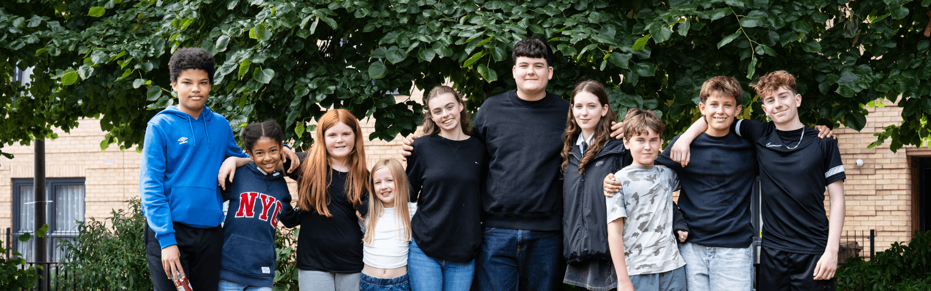 A group of ten young people standing together outdoors in front of a large leafy tree.